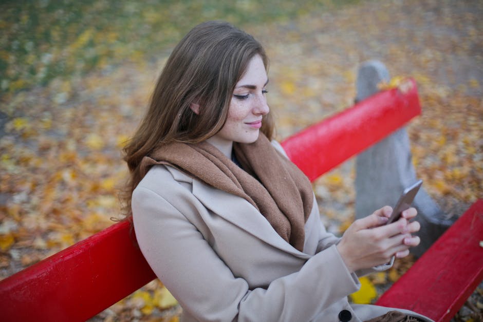 A person strolling along a leaf-covered path in warm autumn light, relaxed posture, glancing down at a phone